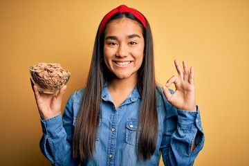 Canvas Print - Beautiful asian woman holding bowl with healthy corn flakes cereals over yellow background doing ok sign with fingers, excellent symbol