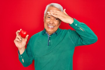 Wall Mural - Middle age senior grey-haired man holding romantic red heart object over isolated red background stressed with hand on head, shocked with shame and surprise face, angry and frustrated. Fear and upset