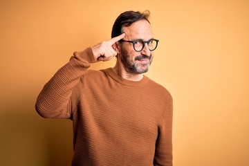 Middle age hoary man wearing brown sweater and glasses over isolated yellow background Smiling pointing to head with one finger, great idea or thought, good memory