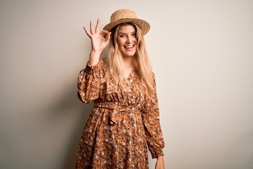 Young beautiful blonde woman wearing summer dress and hat over isolated white background smiling positive doing ok sign with hand and fingers. Successful expression.