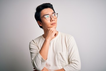 Young handsome chinese man wearing casual t-shirt and glasses over white background with hand on chin thinking about question, pensive expression. Smiling with thoughtful face. Doubt concept.