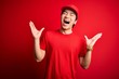 Young handsome chinese delivery man wearing cap standing over isolated red background celebrating mad and crazy for success with arms raised and closed eyes screaming excited. Winner concept