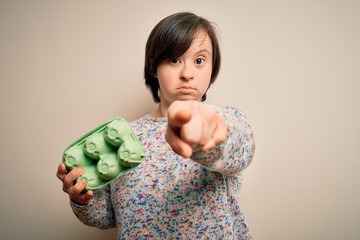 Sticker - Young down syndrome woman holding cardboard egg cup from fresh healthy eggs pointing with finger to the camera and to you, hand sign, positive and confident gesture from the front