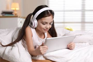 Sticker - Cute little girl with headphones and tablet listening to audiobook in bed at home