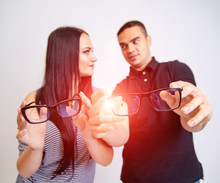 Lovely Attractive Couple Sitting Together And Looking Each Other Hold Glasses In Their Hands. Young Couple Smiling To One Another Show Eyeglasses In Black Frame Sitting On White Background.