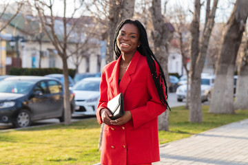 Wall Mural - Close up portrait of a beautiful young african american woman with pigtails in a red business suit smiling and walking along the street