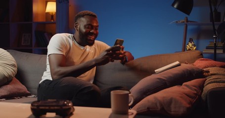 Happy young man laughing and typing message while sitting on sofa at home. Bearded guy smiling while using his smartphone for communication in social networks. Concept of leisure.