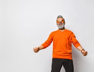 Wall Mural - Elderly man in orange sweater, sunglasses, black pants, bracelets. Raised fists , screaming, posing isolated on white. Close up