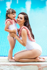 Young Mom with baby daughter in the same white swimsuits sitting on the edge of the pool. Clothes for summer and sea family look.