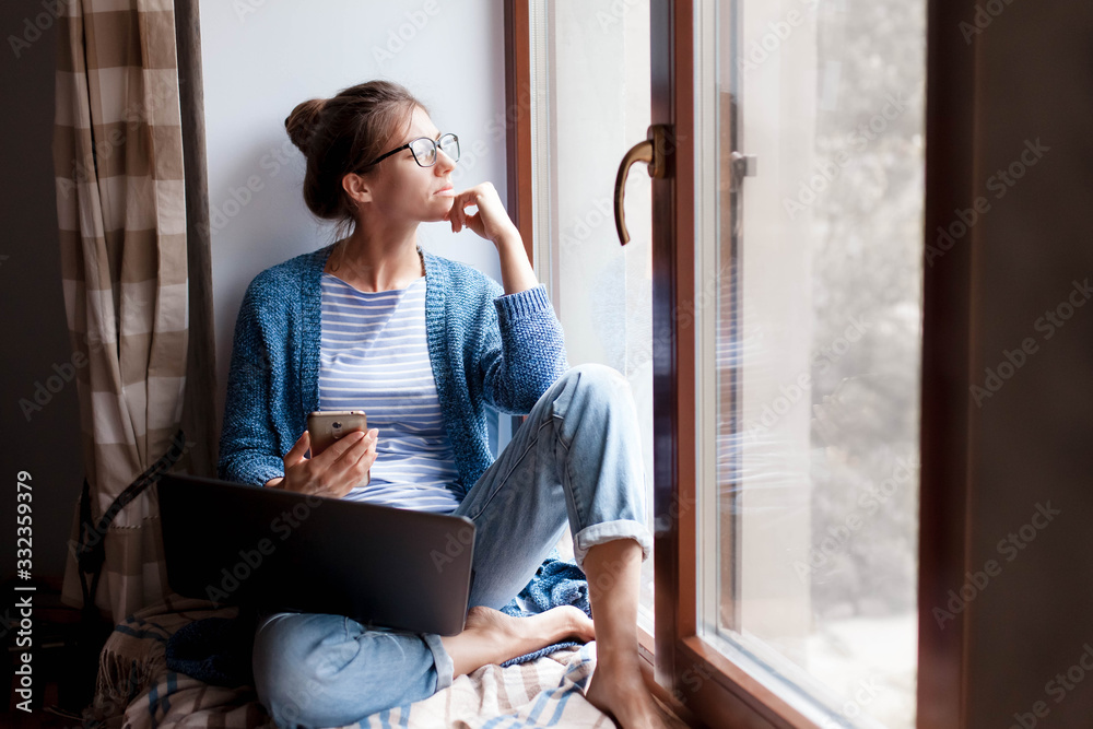 Remote working from home office. Young woman using laptop, phone. Freelancer workplace by window. Teleworking in isolation, female business, shopping online, distance education. Lifestyle moment. - obrazy, fototapety, plakaty 