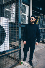 City portrait of handsome hipster guy with beard wearing black blank hoody or sweatshirt and hat with space for your logo or design. Mockup for print