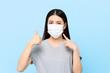 Leinwandbild Motiv Young Asian woman wearing face mask to protect from COVID-19 and giving thumbs up isolated on light blue background
