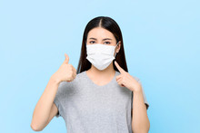 Young Asian Woman Wearing Face Mask To Protect From COVID-19 And Giving Thumbs Up Isolated On Light Blue Background