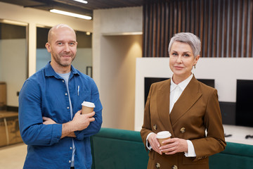 Wall Mural - Waist up portrait of two mature people holding coffee cups standing in modern office interior and smiling at camera, copy space