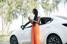 Cheerful young Black woman with long hair standing next to her car and sending text messages to friends