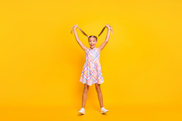 Poster - Full length photo of cheerful beautiful little lady good mood raise pretty long tails up air playful girl wear plaid summer dress sneakers isolated yellow bright color background