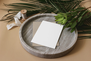 Wall Mural - Summer tropical wedding stationery mock-up scene. Blank greeting card, envelope, ribbon and green palm leaves and leucadendron flowers on marble tray. Beige table background. High angle top view.