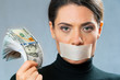 a girl with her mouth closed with a white ribbon holds a lot of dollars in her hand and looks at the camera. silent witness concept, corruption, censorship.