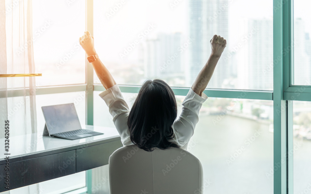 Business achievement concept with happy businesswoman relaxing in office or hotel room, resting and raising fists with ambition looking forward to city building urban scene through glass window - obrazy, fototapety, plakaty 