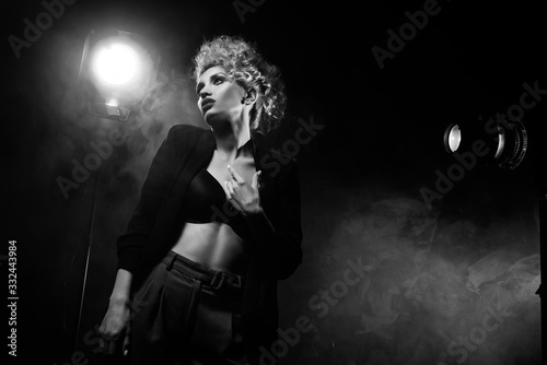 Fotografía A beautiful blonde girl with an elegant hairstyle and large breasts, wearing a bra, trousers and a blazer, artistically poses in the rays of spotlights in the smoke