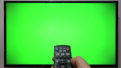 Person pointing a TV remote and pressing buttons in front of a TV with green screen
