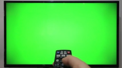 Person pointing a TV remote and pressing buttons in front of a TV with green screen
