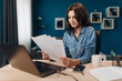 © Kuz Production - Attractive young brunette in casual clothing holding documents while sitting at table with personal laptop. Happy female with curly hair doing paperwork at home.