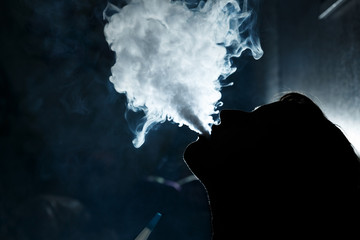 Silhouetted by a girl who smokes a hookah at night in a nightclub. It releases thick smoke from the mouth.