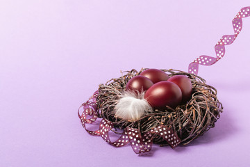 On a pink background is a nest of thin brown branches, decorated with a white bird feather and ribbon. There are three red eggs in it. Easter holiday.