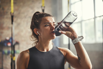 Close up of young athletic woman in earphones is drinking water while exercising in gym.