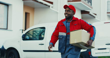 Attractive Young African American Mailman In Red Costume And Cap Taking Out Percel From A Van And Walking To The House To Deliver It. Outdoors.