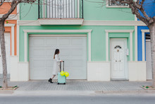 Side View Of Young Stylish Female With Yellow Balloons And Suitcase Walking On City Street Next To Old Styled Colorful Building