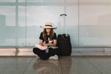 Young Woman In Hat And Casual Wear Sitting In Lotus Pose On Floor Messaging On Smartphone Waiting For Flight In Airport