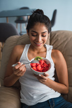 Beautiful Brunette Hispanic Woman Holding Fork And Bowl Of Kiwi And Watermelon And Sitting On Settee