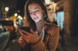 Attractive happy young woman reading good news on her mobile phone during night walk in the city streets with bokeh light on the blurred background, Beautiful smiling girl using smartphone outdoors