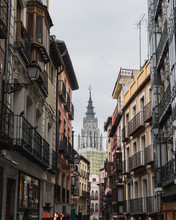 From Below Of Narrow Street Of City Toledo In Spain With Old Residential Buildings And Cathedral In Background Against Gray Cloudy Sky