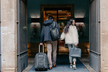 Back View Of Happy Stylish Young Couple In Love With Suitcase And Bags Holding Hands And Entering Modern City Hotel During Travel While Woman Looking At Camera Over Shoulder
