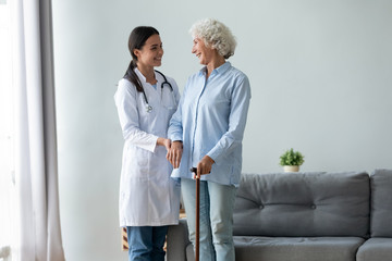 Happy caring young woman doctor or nurse and positive old lady with walking stick stand hugging talking, attentive caregiver support comfort optimistic senior grandmother give help and assistance