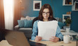 © MYDAYcontent - Focused young woman working at home looking at papers sitting at desk next to laptop computer