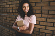 © BullRun - Happy curly dark skinned 20s woman writer enjoying free time outdoors near wall holding planner, attractive african american hipster girl in trendy casual wear posing with book looking away
