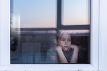 A Sad, Lonely Child Sits On The Windowsill And Looks Out The Window. Stay At Home Quarantine Coronavirus Prevention Of The Pandemic. The View From The Street. Prevention Of The Epidemic.
