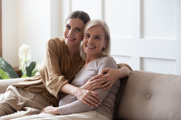Wall Mural - portrait happy adult daughter and older mother hugging and holding hands, sitting on couch at home, 