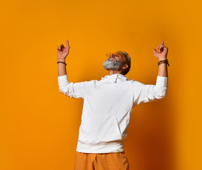 Wall Mural - Elderly male in white hoodie, pants and sunglasses, bracelets. Raised hands up, smiling, posing on orange background. Close up