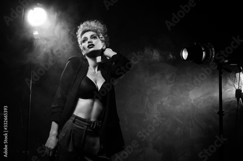 Fotografía A beautiful blonde girl with an elegant hairstyle and large breasts, wearing a bra, trousers and a blazer, artistically poses in the rays of spotlights in the smoke
