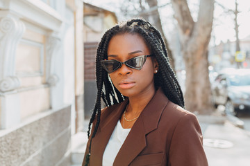 Wall Mural - Close up portrait of a beautiful young african american woman with pigtails hairstyle in a brown business suit walks along spring streets