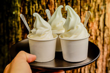 Hand Holding Three Vanilla Ice Scream Paper Cups Ready To Serve On Black Tray With Plastic Spoon On Wood Background. Close Up Milky Soft Cream So Yummy.