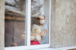 Leinwandbild Motiv Sad Child, little girl in mask with teddy bear looking from window, coronavirus quarantine