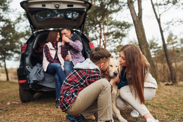 Wall Mural - Happy family sitting and having fun with their dog near modern car outdoors in forest