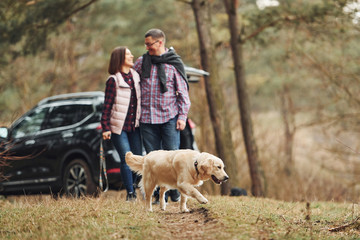 Wall Mural - Happy mature couple have a walk with their dog in autumn or spring forest near modern car