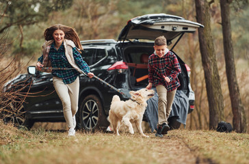 Wall Mural - Cheerful girl with brother have walk with their dog outdoors in forest at autumn or spring season near car